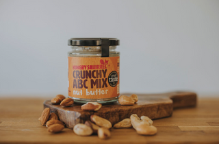 Jar of crunchy nut butter on a wooden board with nuts around, against a plain background
