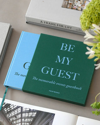 Green guestbook titled 'Be My Guest' on a surface with other books and a plant.