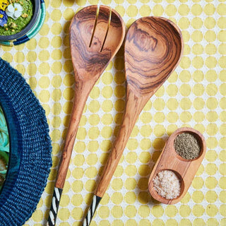 Wooden salad servers with black and white handles beside a small wooden dish with spices on a yellow patterned tablecloth.