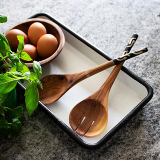 Wooden salad server spoons with black and white handles on a tray with eggs and basil leaves on a stone worktop