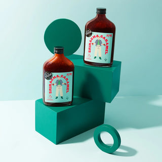 Two glass bottles of Sriracha Caramel on a teal geometric shelf with a light blue background