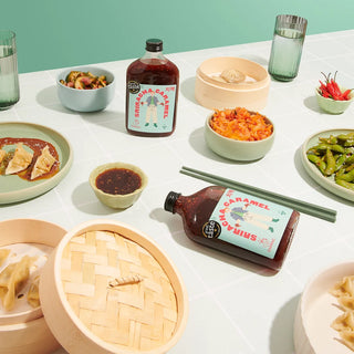 Table with Eastern Asian food and bottles of Chimac Sriracha Caramel sauce against a light green background