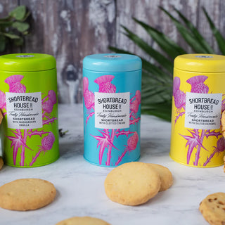 Three colorful tins from Shortbread House of Edinburgh on a marble surface with shortbread rounds beside.