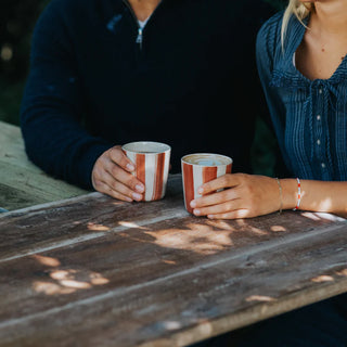 Two people holding striped mugs on a wooden table outdoors.