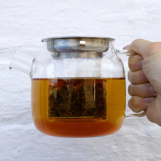 Glass teapot with a handle filled with tea, held by a hand against a white background