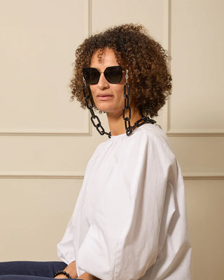 Person wearing a white top and sunglasses with black glasses chain straps against a beige wall.
