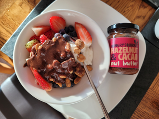Bowl of yogurt with fruit and a jar of hazelnut and cacao nut butter on a wooden table.
