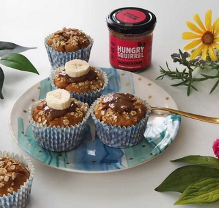 Muffins on a plate with a jar of 'Hungry Squirrel Hazelnut Cacao ' nut butter, surrounded by flowers and leaves.