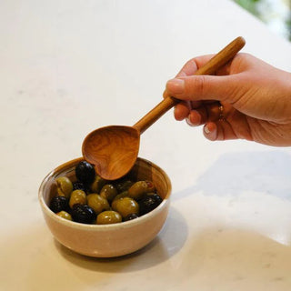 Hand holding a heart shaped wooden spoon over a bowl of olives on a light surface