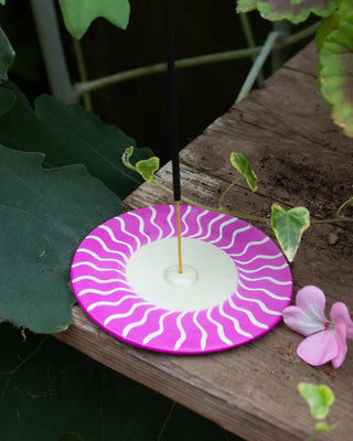 Pink and white striped incense holder with an ivy stem on a wooden surface with leaves and flowers in the background