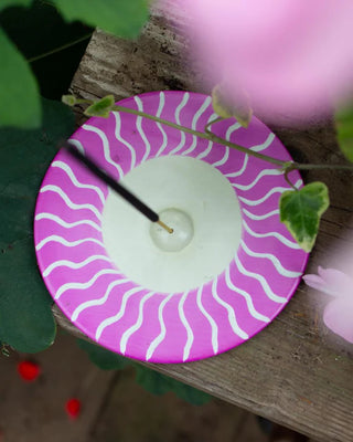 Pink and white wavy-patterned incense holder on a wooden surface with green leaves and pink flowers in the background.