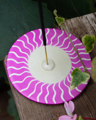 Round Incense holder with pink and white wavy design on a wooden surface with green leaves.