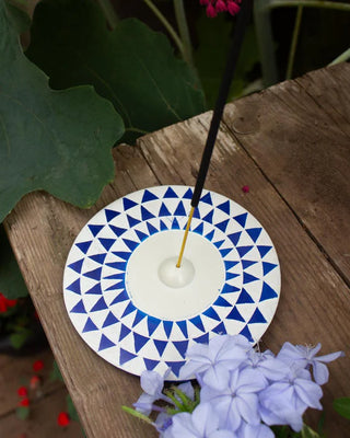 Incense stick on a blue and white round geometric patterned holder with flowers and leaves in the background