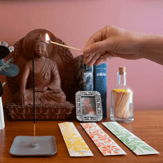Person lighting incense with a match, surrounded by a Buddha statue, books, and a bottle of matches on a wooden table.
