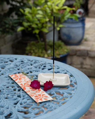 Incense stick on a small white dish with flowers and card packaging on a blue table outdoors.