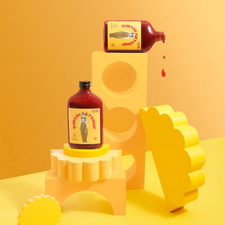 Two glass bottles of kimchi ketchup, one sideways and pouring, on yellow geometric shapes with a yellow background