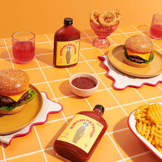 Two burgers with glass ketchup bottles with a yellow label and red font beside fries, and drinks on a yellow checkered tablecloth.