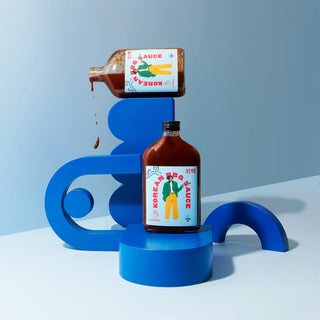 Two glass bottles of Korean BBQ sauce sitting a top blue geometric shapes with one bottle spilling over against a blue background