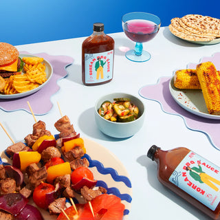 Two glass bottles of Korean BBQ Sauce on a table surrounded by various bbq foods with a pale blue table cloth and darker blue wall