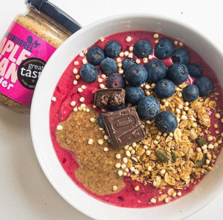 Berry smoothie bowl with granola, blueberries, and nut butter and a jar of Maple Pecan nut butter on a white surface.