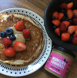 Pancakes with berries and a jar of 'Hungry Squirrel Maple Pecan Nut Butter' spread on a wooden table.