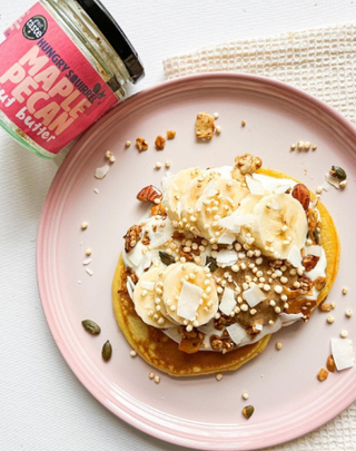 Pink plate with pancakes topped with bananas, nuts and nut butter, next to a jar of maple pecan nut butter.