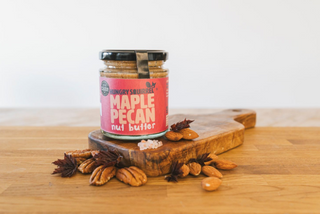 Jar of Maple Pecan nut butter on a wooden cutting board with pecans around it.