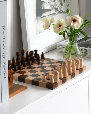 Wooden chess set with pieces on a checkered board, next to a vase of flowers and books.