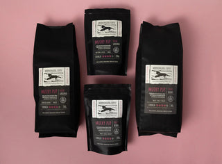 Multiple bags of Mucky Pup coffee pouches set against a pink background