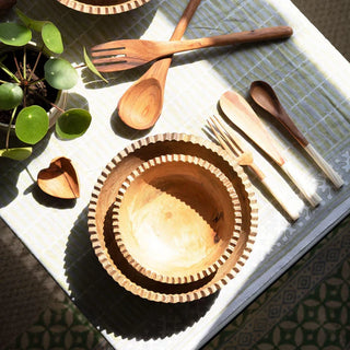 Wooden salad servers set on a table beside wooen bowls & cutlery with a plant and decorative items.