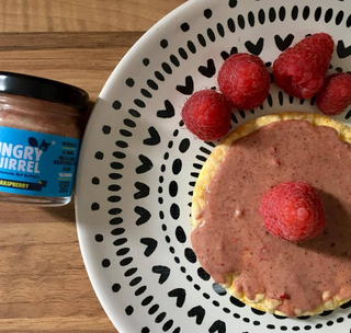 Plate with a pancake topped with Royal Raspberry Almond Butter and raspberries, next to a jar of 'Hungry Squirrel' royal raspberry spread on a wooden surface.