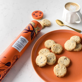 Orange plate with shortbread rounds, a mug of coffee, and a cylindrical tin of Shortbread House cookies on a white surface.