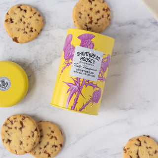 Open Yellow shortbread tin with purple thistle design and 'Shortbread House' label on a marble surface surrounded by shortbread rounds.