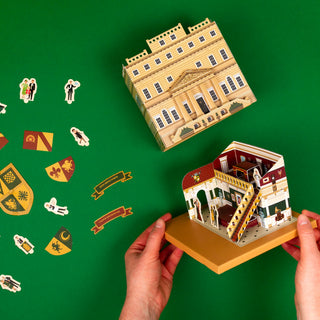 Hands assembling a miniature architectural model on a green background with flat pieces to the side