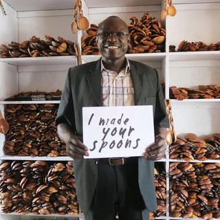 Man holding a sign saying 'I made your spoons' in a workshop setting with shelves of wooden spoons.
