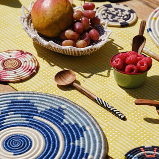 Colorful woven coasters and bowls with fruits and a wooden spoon with black and white handle on a yellow textured surface