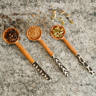 Three wooden spoons with black and white uniquely decorated handles, each filled with different spices on a textured surface.