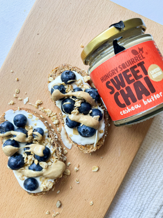 Two crackers with blueberries and cashew butter next to a jar of 'Sweet Chai' cashew butter on a wooden board.