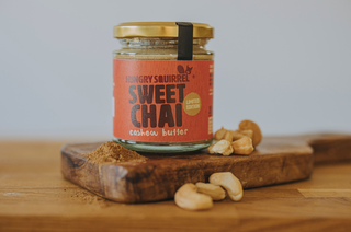 Jar of 'Sweet Chai' limited edition cashew butter on a wooden board with scattered cashews.