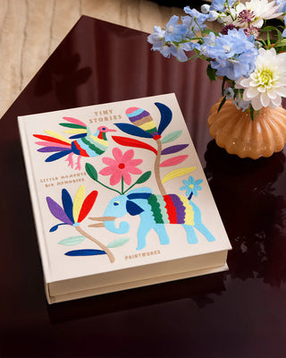 Beige photo album book with multicolour embroidery depicting an elephant, bird and flowers on a dark coffee table beside a vase of flowers